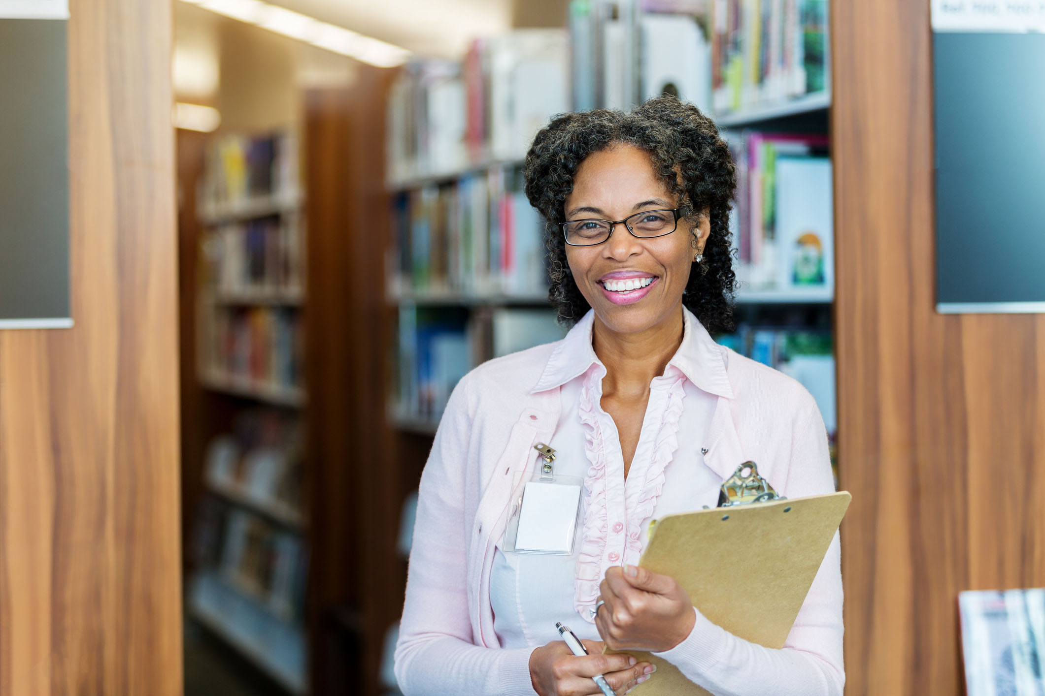 A cheerful mature female school library volunteer smiles for the camera. She is standing in front of a book shelf holding a clipboard and pen and wearing a nametag.
