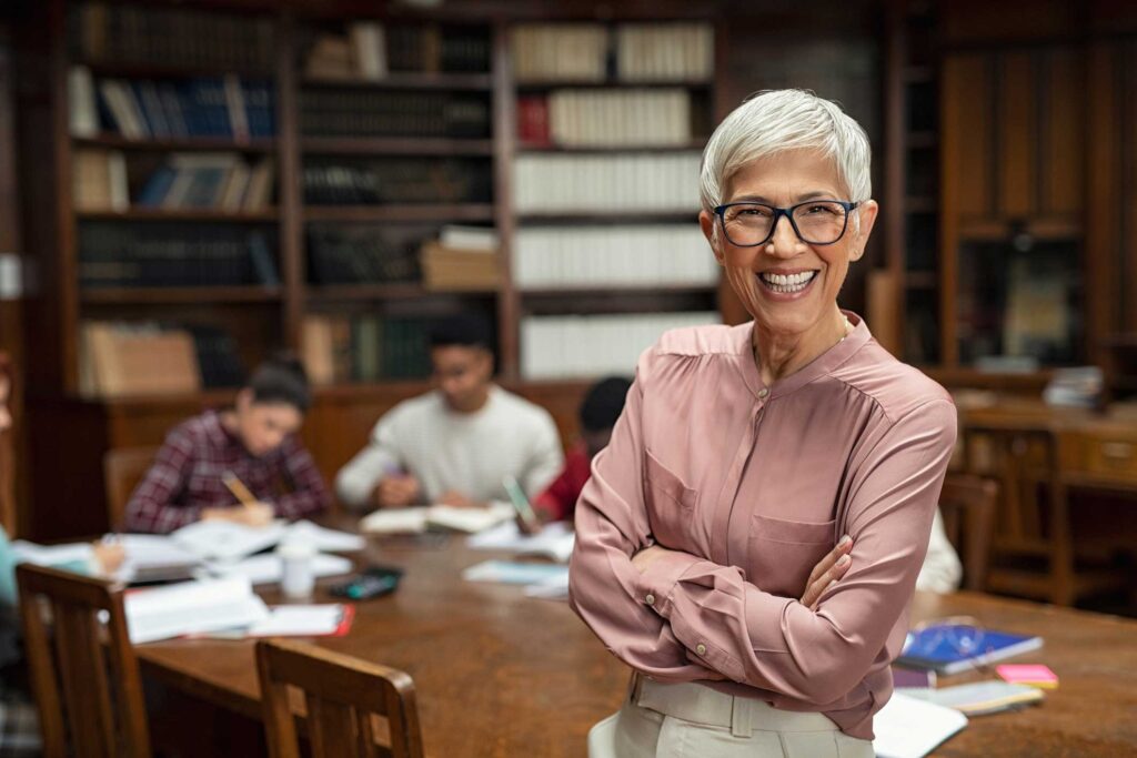 Portrait of mature professor with crossed arms standing in university library and looking at camera with copy space. Happy senior woman at the library working as a librarian. Satisfied college teacher smiling with students in background studying.