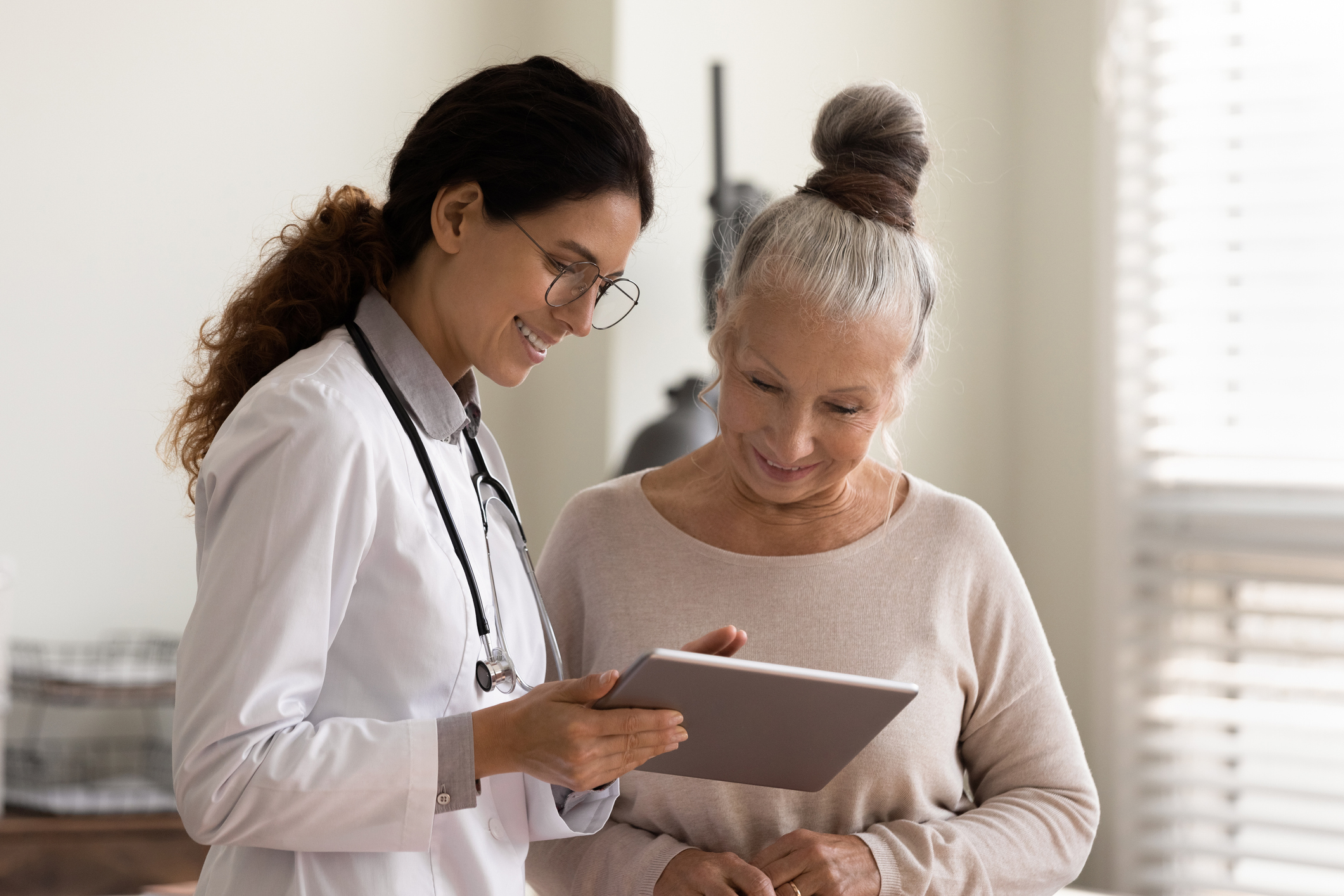 Happy female doctor and senior patient look at tablet screen discuss treatment or therapy on gadget. Smiling woman nurse and mature client use pad device discussing results in modern hospital.